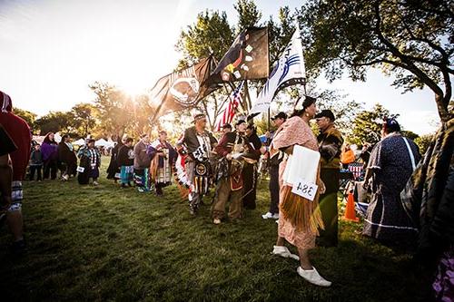 A line of Powwow attendees, some holding large flags representing different tribes.